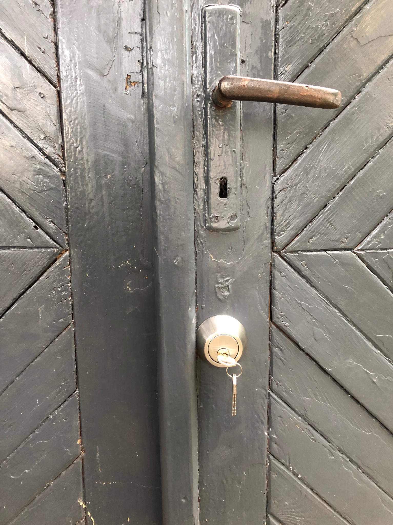 Szczegółowe ujęcie pomalowanych na szaro drzwi z wzorem w jodełkę, z zamontowanym nowym zamkiem cylindrycznym, w którym tkwią klucze, oraz starą, rdzewiejącą klamką.