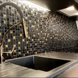 Czarny, granitowy zlew kuchenny z baterią w kolorze polerowanego złota na tle ściany wyłożonej czarno-złotą mozaiką w małe kwadraty, podświetloną listwą LED.