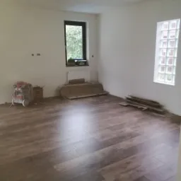 Pokój po położeniu paneli podłogowych, widoczny sprzęt budowlany, okno i fragment grzejnika