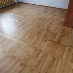 Jasny parkiet ułożony w wzór prostokątów z wstawkami, widoczny grzejnik i fragment ściany