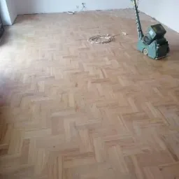 Cyklinowanie parkietu w jodełkę: maszyna do cyklinowania stoi na częściowo odnowionej podłodze, widoczny kabel zasilający, ściany w trakcie remontu.