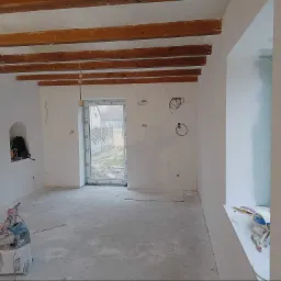 Wnętrze w trakcie remontu: surowe ściany, drewniane belki stropowe, przygotowany odkurzacz przemysłowy i akcesoria malarskie na parapecie okiennym.