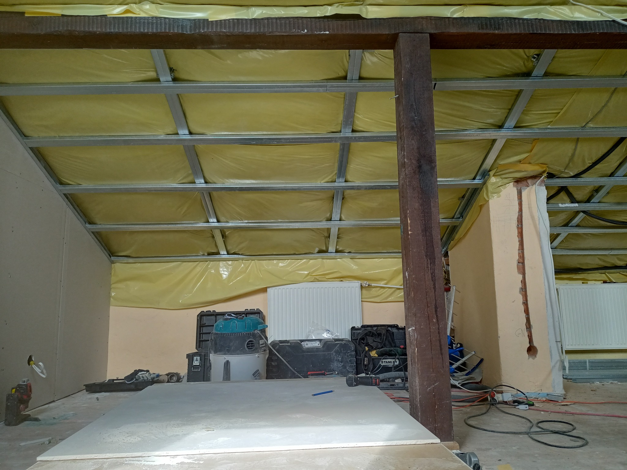 Wnętrze w trakcie remontu z widoczną konstrukcją metalową sufitu podwieszanego, izolacją z folii, narzędziami i płytą gipsowo-kartonową na podłodze.
