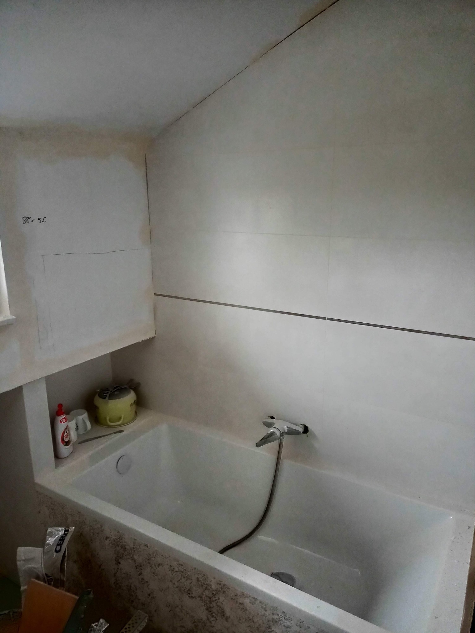 Łazienka w trakcie remontu: biała wanna, ściany częściowo wyłożone jasnymi płytkami, widoczne ślady po wcześniejszej instalacji, zaznaczone ołówkiem obszary na ścianie, narzędzia i chemia budowlana...