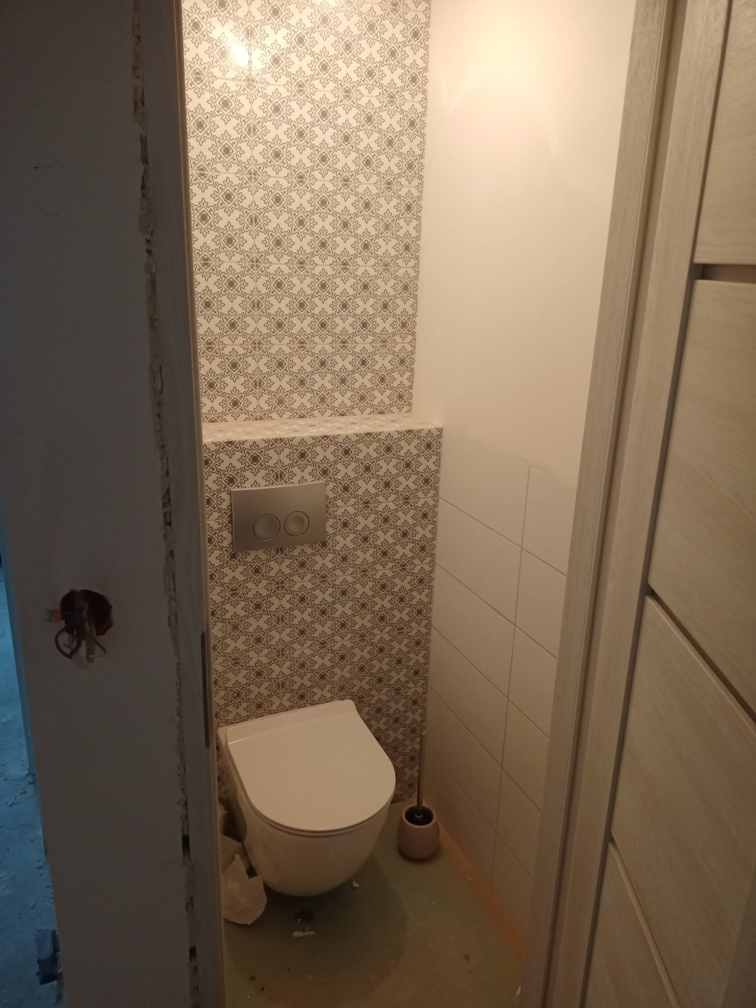 Toaleta wisząca w małej łazience z częściowo wykończonymi ścianami: dekoracyjne płytki za toaletą i białe płytki po prawej stronie, widok z korytarza przez niedokończone ościeże drzwi.