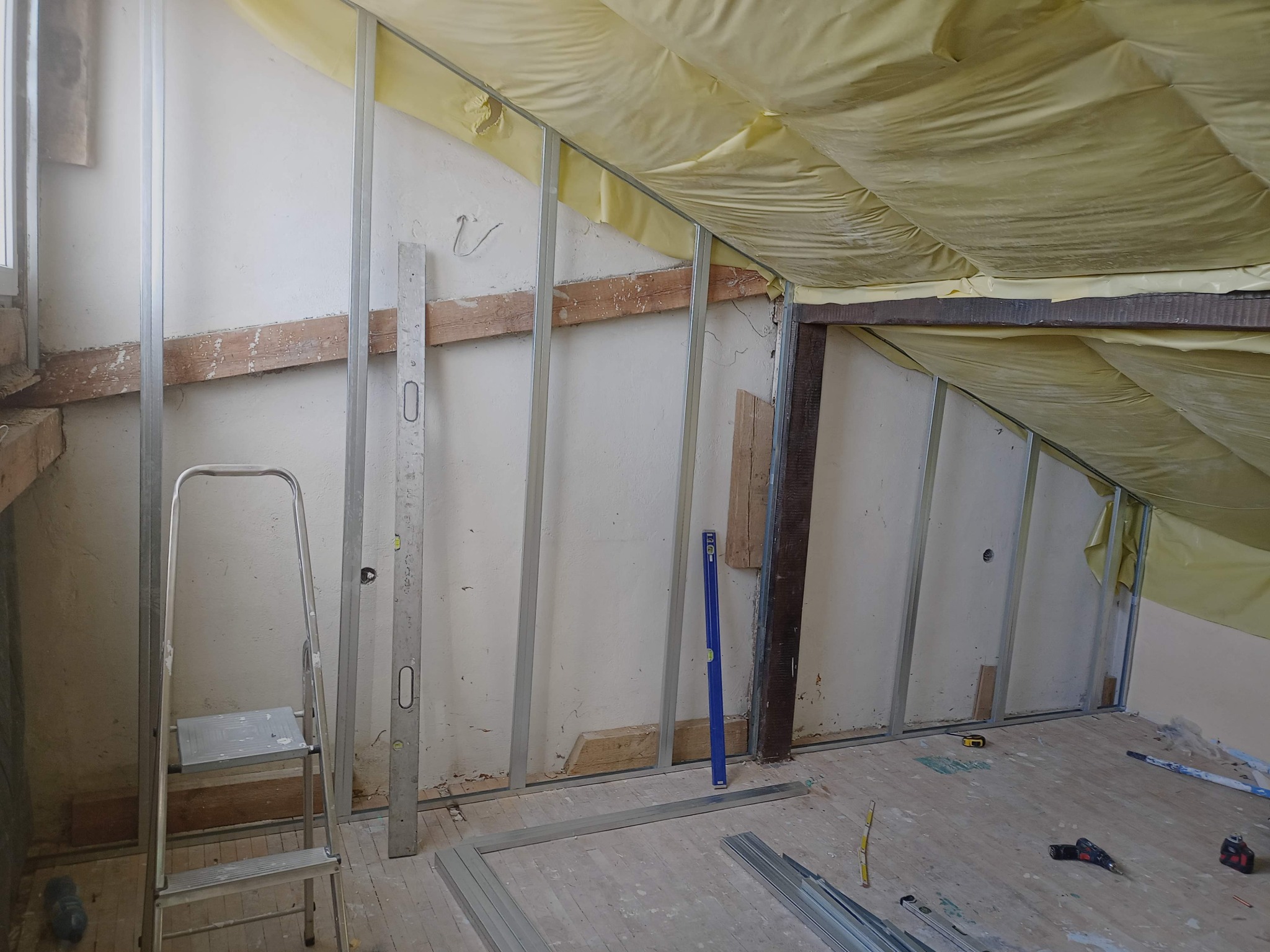 Montaż metalowego stelaża pod płyty gipsowo-kartonowe na poddaszu ze skośnym dachem, widoczna izolacja z żółtej folii, poziomice, drabina aluminiowa i narzędzia na podłodze z desek.