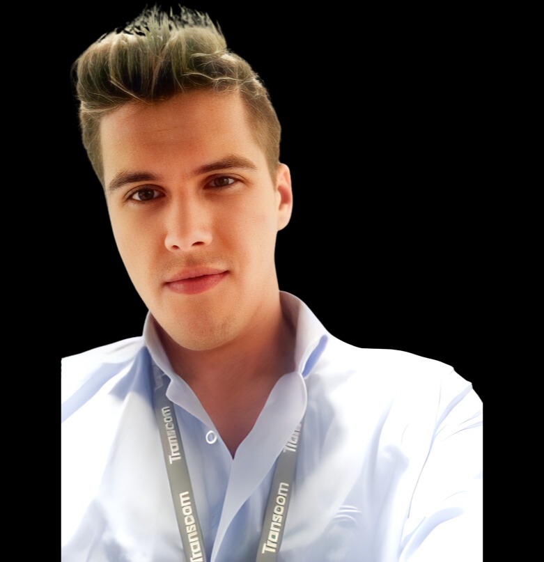 Portret młodego mężczyzny w błękitnej koszuli z identyfikatorem Transcom na smyczy, uśmiechającego się do kamery na czarnym tle.