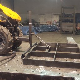 Usługi Spawalnicze Jakub Bujak - Rama metalowa z podziałem na kwadraty, zamocowana do żółtego traktorka jednoosiowego na stole spawalniczym w warsztacie, w tle wózek widłowy i elementy konstrukcji stalowych.