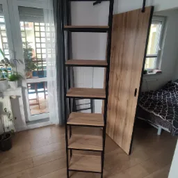 Wysoki, metalowy regał z drewnianymi półkami, stojący na drewnianej podłodze w pomieszczeniu z oknem balkonowym i przesuwnymi drzwiami.