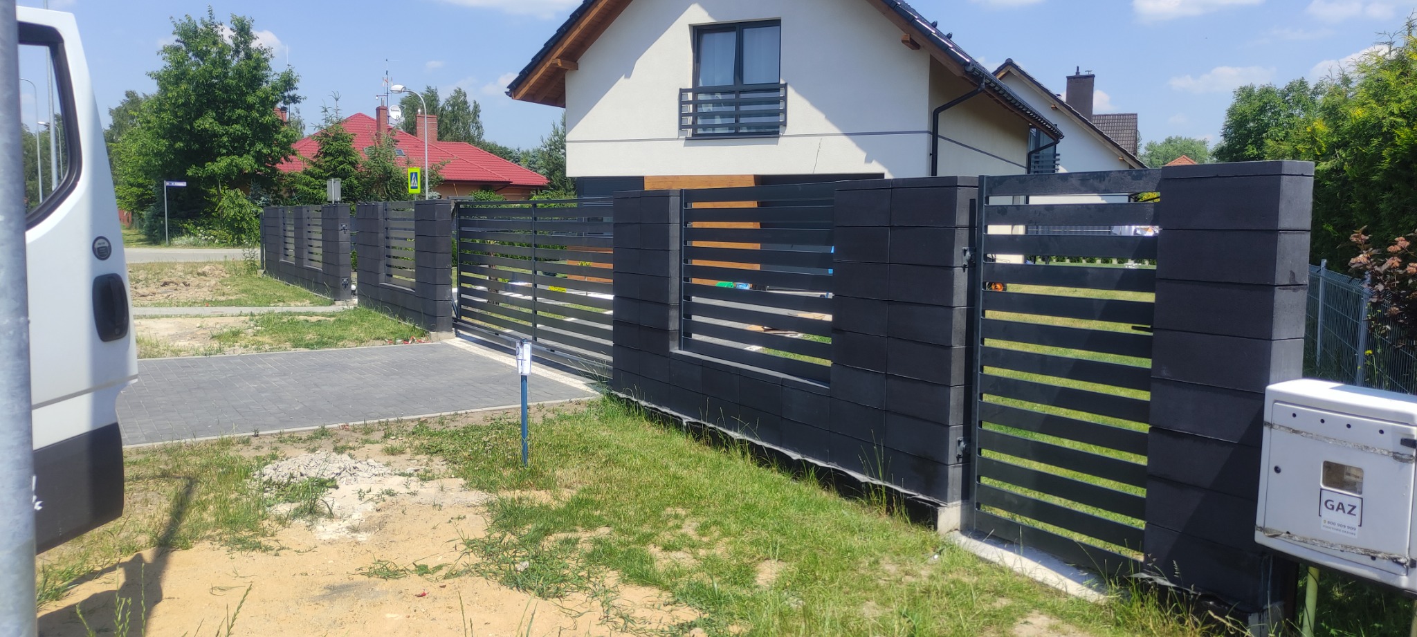 Nowoczesne, grafitowe ogrodzenie panelowe z betonowymi słupkami, przesuwna brama wjazdowa, widok z zewnątrz na tle domu jednorodzinnego i skrzynki gazowej.