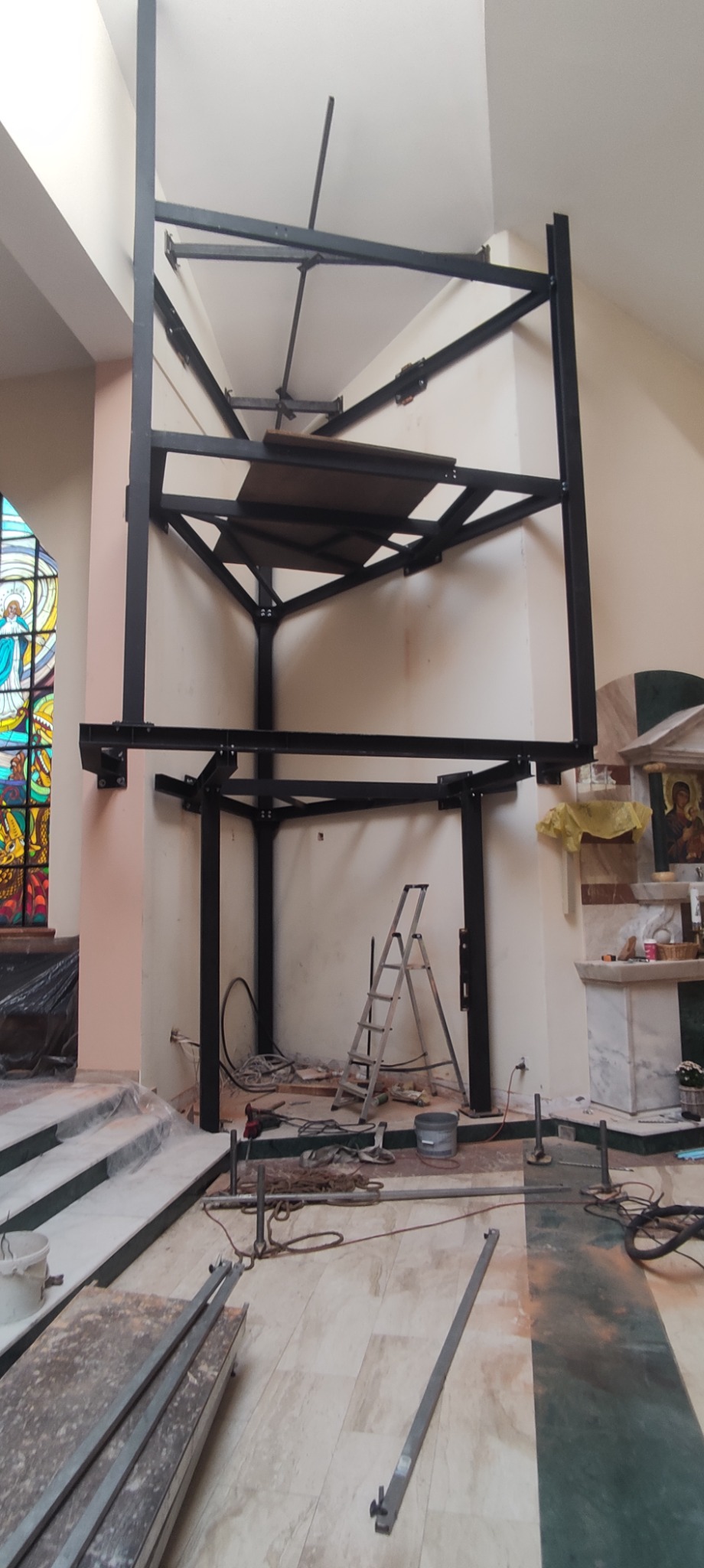 Wewnętrzna stalowa konstrukcja w trakcie montażu w kościele, widoczne elementy metalowe, drabina, narzędzia i fragment witrażu.