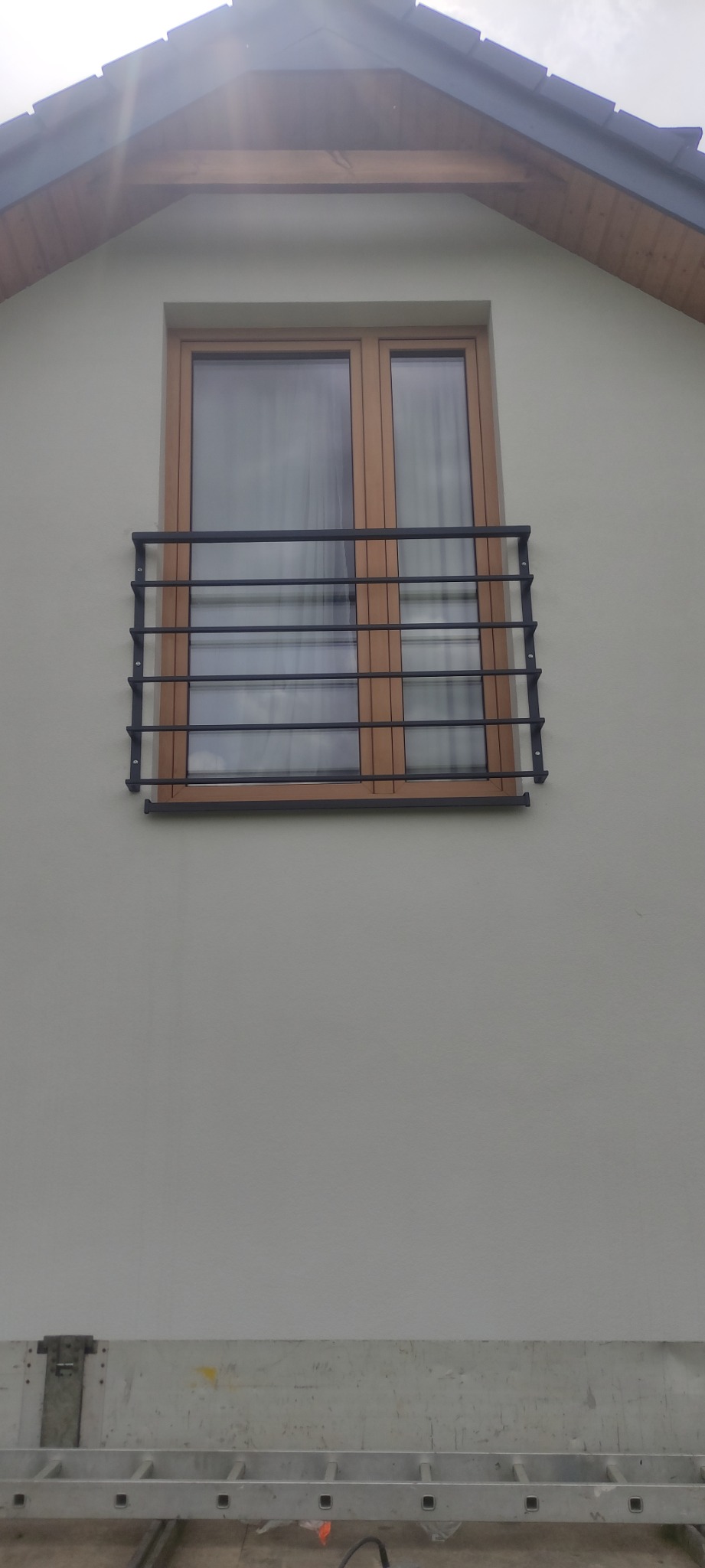 Metalowa balustrada okienna z poziomymi prętami, zamontowana na elewacji budynku z drewnianą ramą okienną, widok z dołu na tle drabiny.