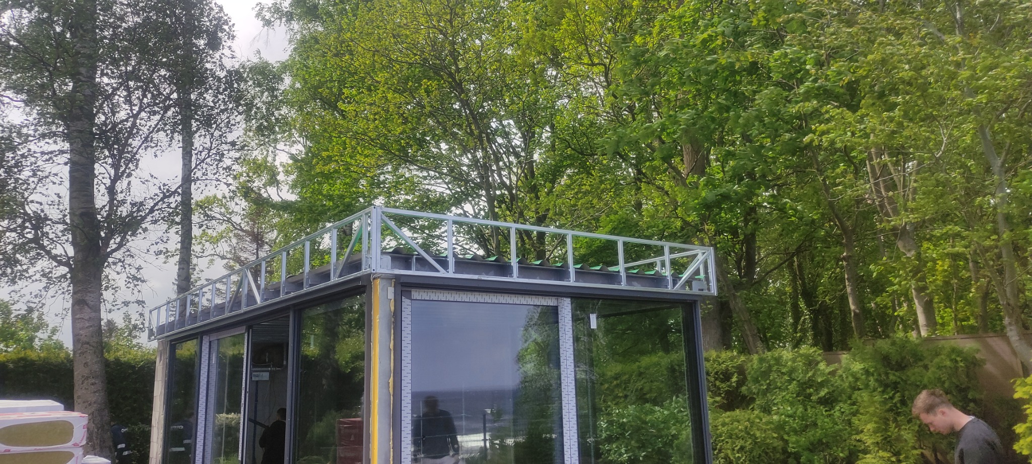 Szklana konstrukcja budynku z metalowym szkieletem dachu w otoczeniu drzew, widoczne materiały izolacyjne i sylwetki osób wewnątrz.