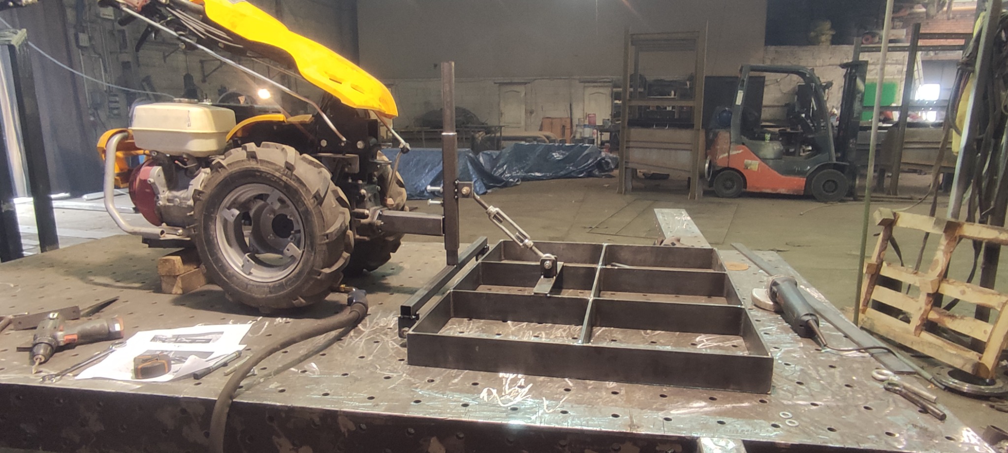 Rama metalowa z podziałem na kwadraty, zamocowana do żółtego traktorka jednoosiowego na stole spawalniczym w warsztacie, w tle wózek widłowy i elementy konstrukcji stalowych.
