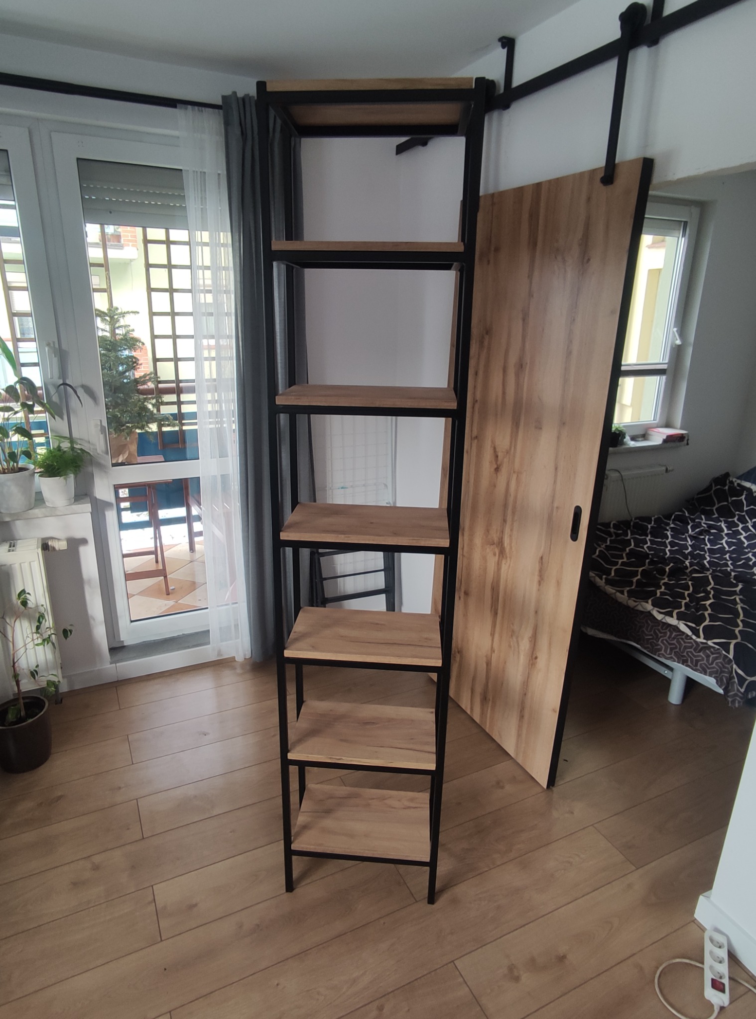 Wysoki, metalowy regał z drewnianymi półkami, stojący na drewnianej podłodze w pomieszczeniu z oknem balkonowym i przesuwnymi drzwiami.