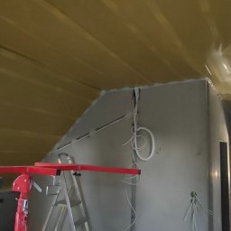 Klein bau - Montaż płyt karton gips na poddaszu: widoczna konstrukcja z profili, częściowo zaizolowany strop folią, podnośnik do płyt i drabina, instalacja elektryczna w ścianie, w tle sylwetka osoby.