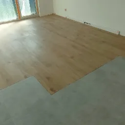 Fragment pokoju z częściowo ułożoną podłogą z paneli drewnopodobnych i płytek ceramicznych, widoczne narzędzia glazurnicze i pomarańczowy element dystansowy.