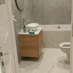 Nowoczesna łazienka w odcieniach szarości i bieli z drewnianą szafką pod umywalką, okrągłym lustrem z podświetleniem LED i marmurową podłogą, widok zza uchylonych drzwi.