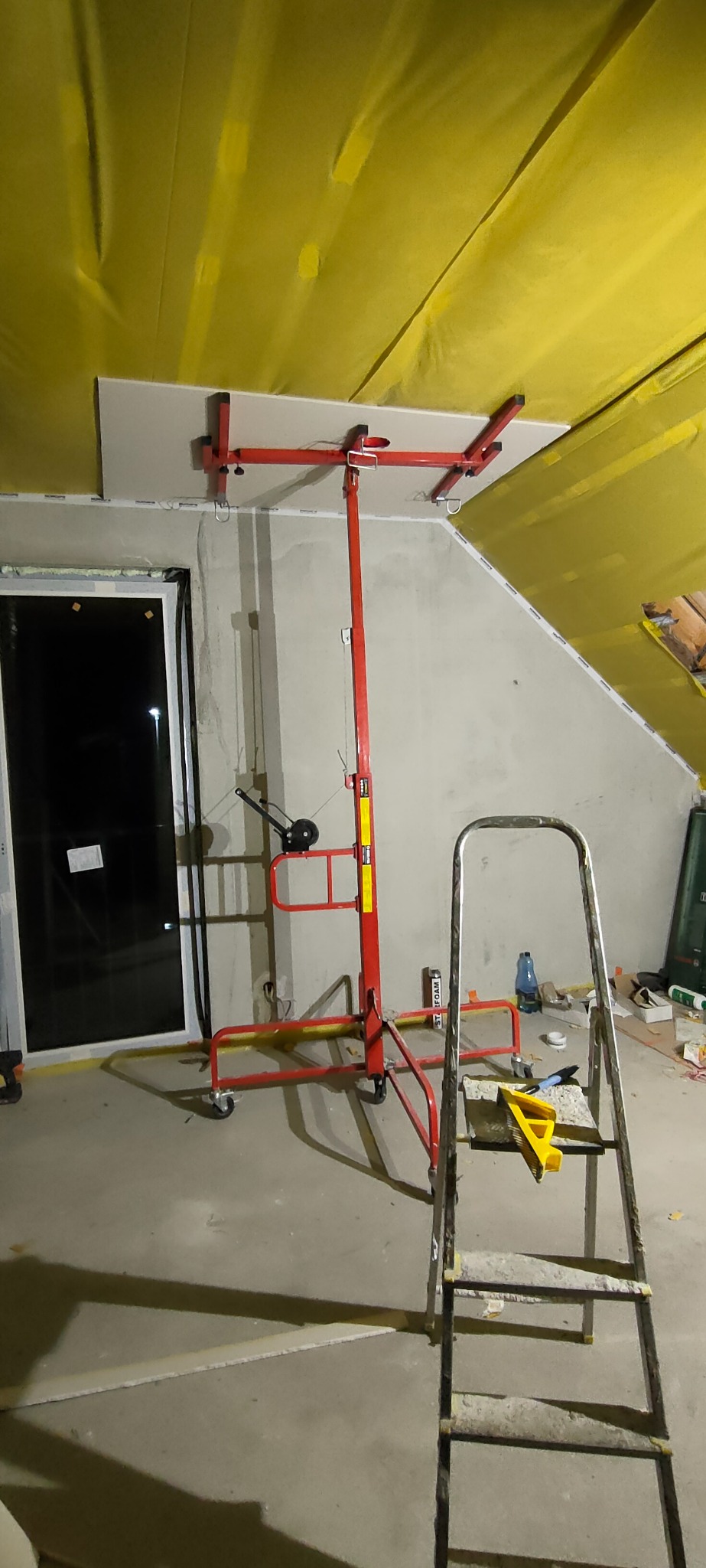 Montaż płyty gipsowo-kartonowej na poddaszu z użyciem podnośnika, widoczna drabina i folia izolacyjna na skosach dachu.