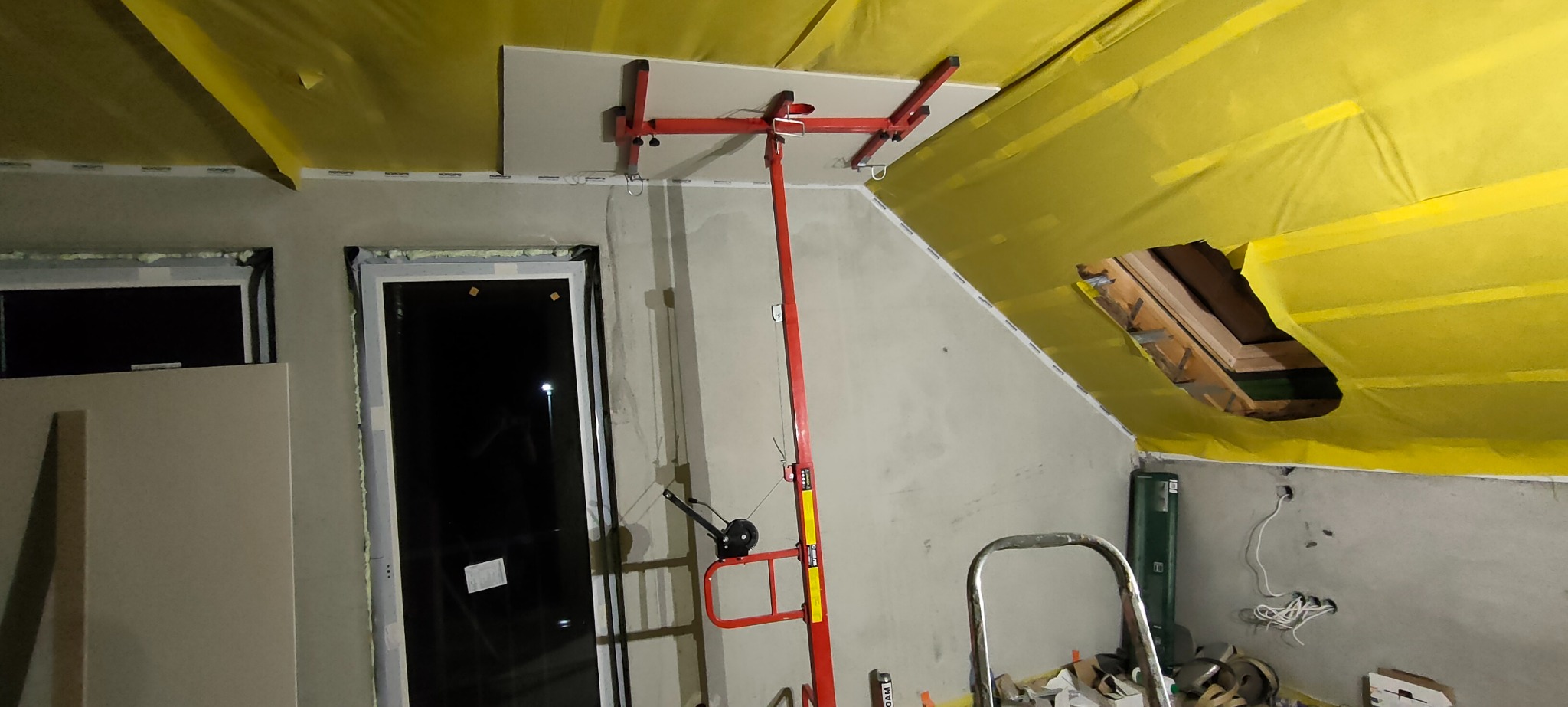 Montaż płyty na poddaszu z oknem dachowym, widoczna żółta folia izolacyjna, podnośnik do płyt w kolorze czerwonym, okna wstawione