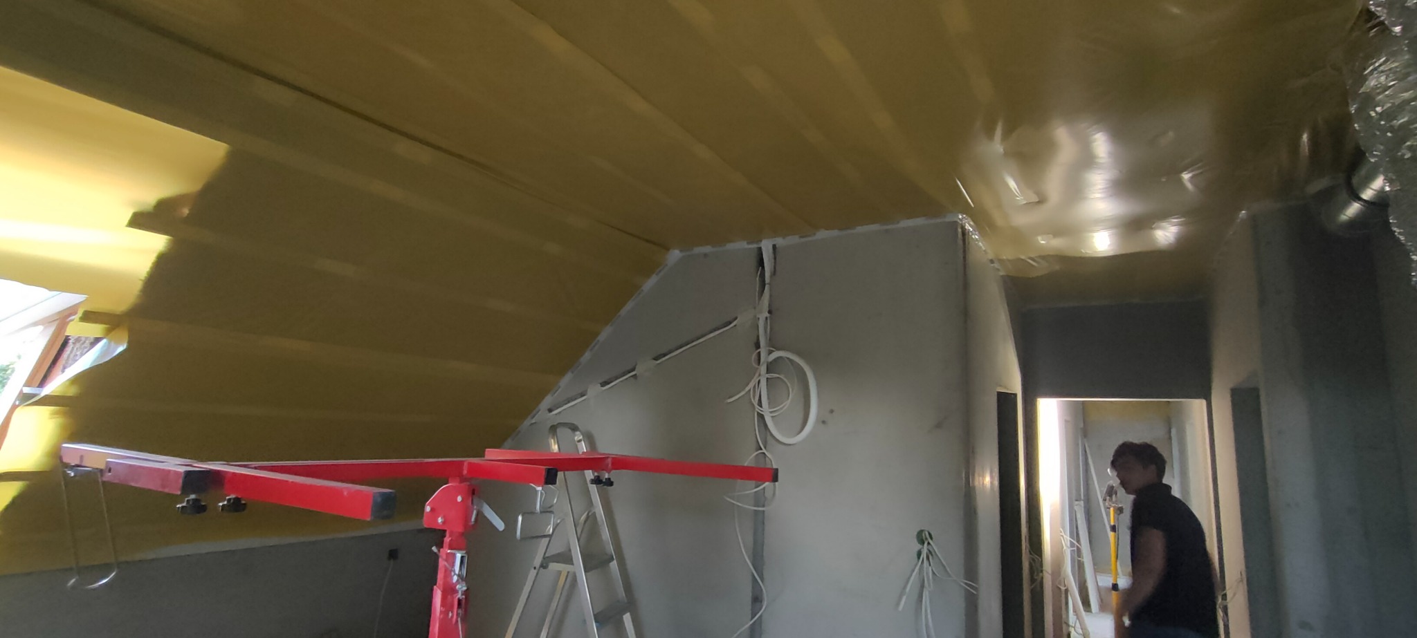Montaż płyt karton gips na poddaszu: widoczna konstrukcja z profili, częściowo zaizolowany strop folią, podnośnik do płyt i drabina, instalacja elektryczna w ścianie, w tle sylwetka osoby.