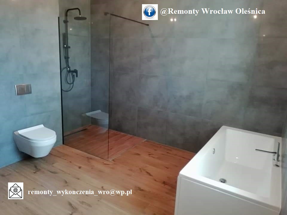 Nowoczesna łazienka z szarą płytką, drewnianą podłogą, kabiną prysznicową typu walk-in i podwieszaną toaletą oraz białą wanną, widoczne logo firmy na zdjęciu.