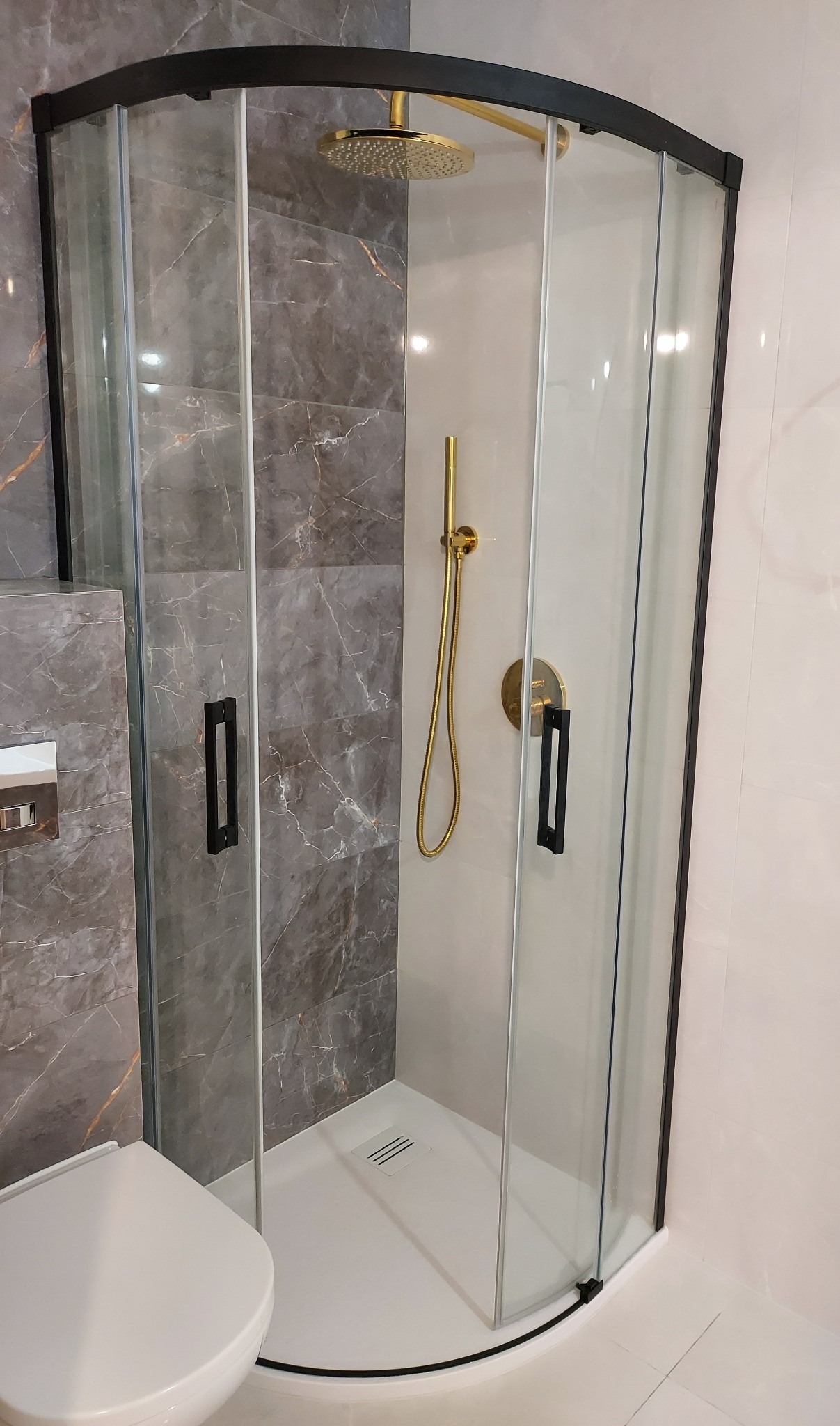 Nowoczesna kabina prysznicowa narożna z czarnymi profilami i złotą armaturą, marmurowe płytki na ścianie, biały brodzik, widoczny fragment toalety.