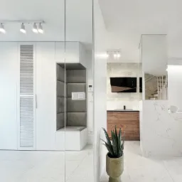 Nowoczesny przedpokój z lustrzaną szafą, wbudowanym siedziskiem z szarą tapicerką i widokiem na kuchnię z drewnianymi szafkami oraz marmurowym blatem, uzupełniony rośliną doniczkową w ceramicznej...