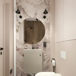 Kompaktowa łazienka w odcieniach beżu z motywem liści na tapecie, okrągłe lustro podświetlone czarnymi lampkami, minimalistyczna umywalka i toaleta.
