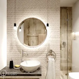 Nowoczesna łazienka z beżowymi płytkami ułożonymi w jodełkę, okrągłym lustrem podświetlonym od tyłu, wiszącymi lampami z żarówkami Edisona, białą umywalką nablatową i szklanym prysznicem.