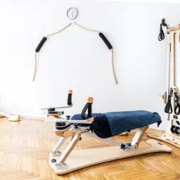 Jasne wnętrze gabinetu z drewnianą podłogą w jodełkę, wyposażonego w specjalistyczne, drewniane urządzenia do ćwiczeń, w tym reformer z niebieskim ręcznikiem, oraz zegar na ścianie.