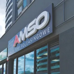 Zewnętrzny szyld firmy 'AMSO Komputery Poleasingowe' z logo i informacją o autoryzacji Microsoft, umieszczony nad witryną sklepu z widocznym odbiciem chmur i budynków w szybach.