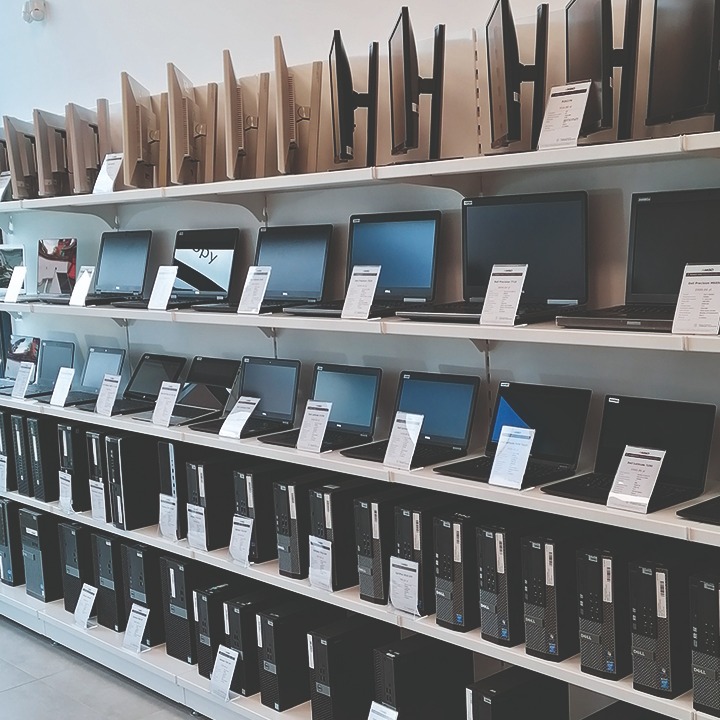Rzędy komputerów stacjonarnych, laptopów i monitorów Dell na półkach w sklepie, z etykietami cenowymi.