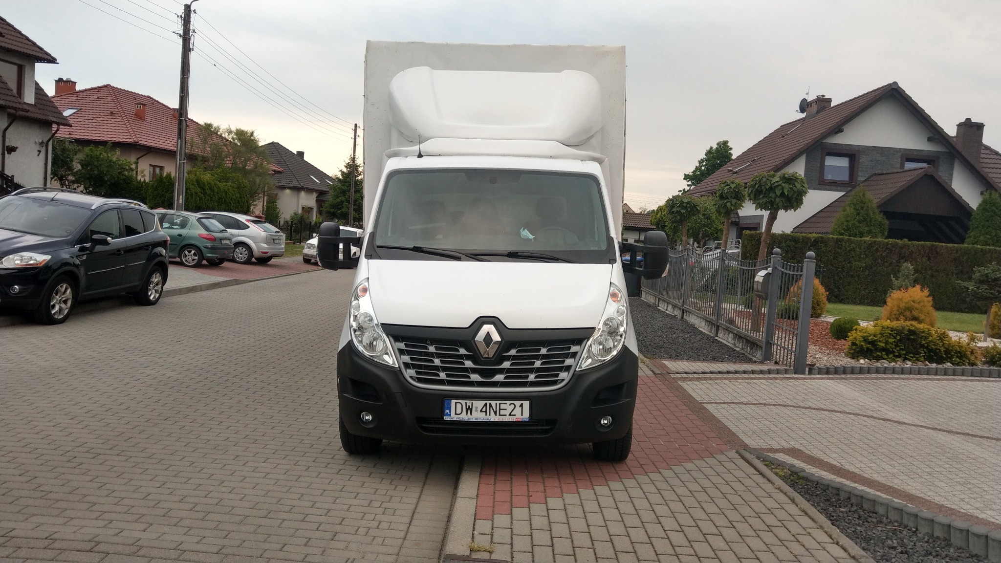 Biały samochód dostawczy Renault Master z zabudową kontenerową na tle domów jednorodzinnych, zaparkowany na brukowanej ulicy. Widoczna tablica rejestracyjna DW 4NE21.
