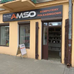 Fasada budynku firmy AMSO z szyldem 'Microsoft Registered Refurbisher', 'AMSO Komputery Poleasingowe', okno z kratą, stojak reklamowy AMSO i widoczne wnętrze sklepu z p&oacute;łkami pełnymi komputer&oacute;w.