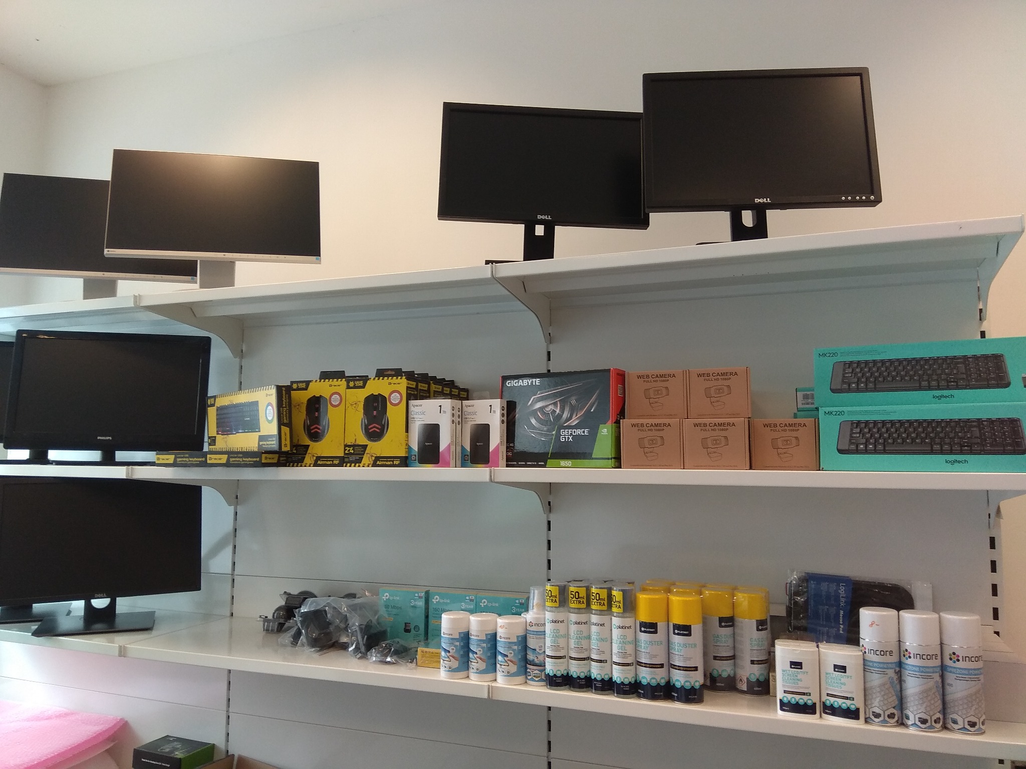 Ekspozycja monitorów, klawiatur, myszek i akcesoriów komputerowych na dwóch białych półkach w sklepie. Widoczne produkty marek Dell, Philips, Logitech, Gigabyte, Apacer i Platinet.