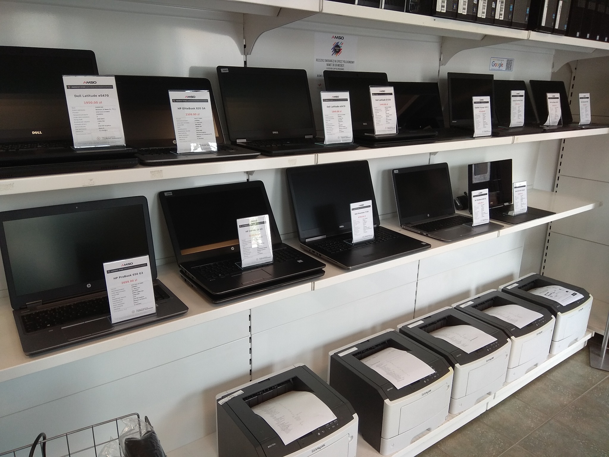 Ekspozycja laptopów i drukarek Lexmark na białych półkach sklepowych z widocznymi cenami na etykietach, zróżnicowane modele i konfiguracje.