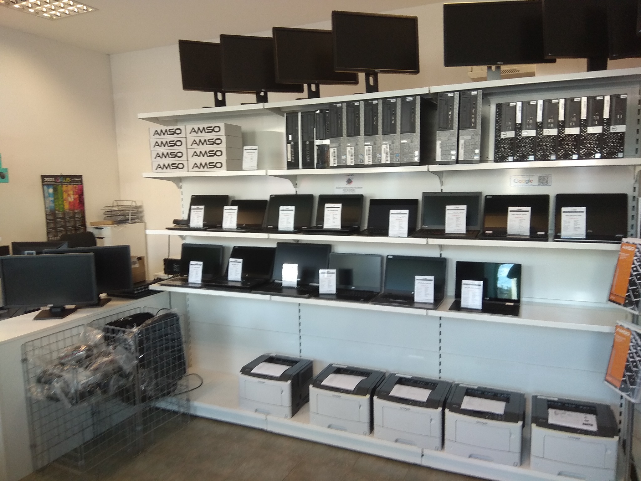 Sklep z elektroniką: rzędy monitorów, komputerów stacjonarnych, laptopów i drukarek na białych półkach, z pudełkami 'AMSO' ułożonymi w stos na górnej półce.