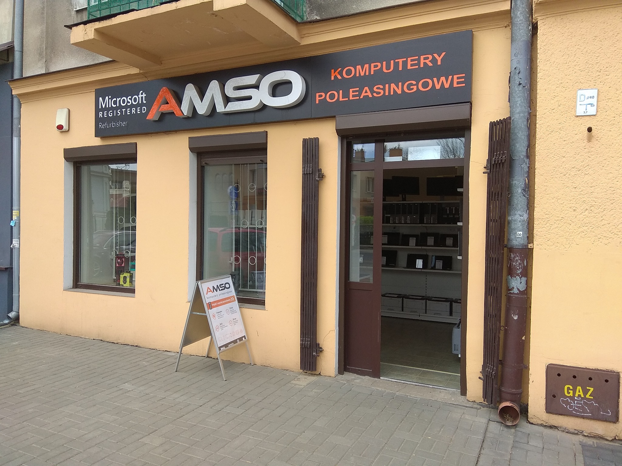 Fasada budynku firmy AMSO z szyldem 'Microsoft Registered Refurbisher', 'AMSO Komputery Poleasingowe', okno z kratą, stojak reklamowy AMSO i widoczne wnętrze sklepu z półkami pełnymi komputerów.