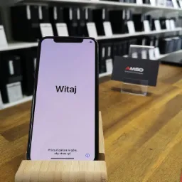 Smartfon iPhone na drewnianym stojaku z ekranem powitalnym 'Witaj', obok wizytówka firmy AMSO na biurku, w tle regał z elektroniką.