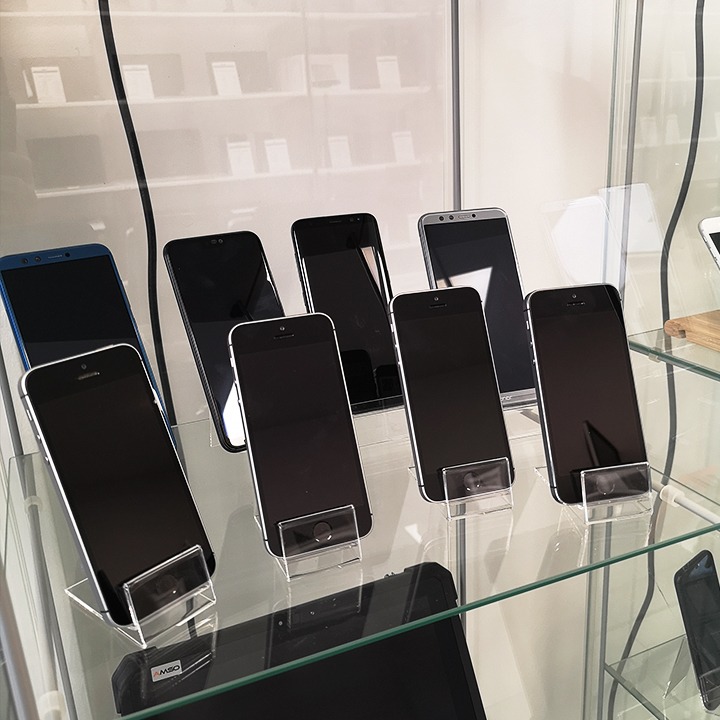Siedem smartfonów różnych marek i modeli, ustawionych na przezroczystych podstawkach w szklanej gablocie, z widocznym tabletem w dolnej części kadru.