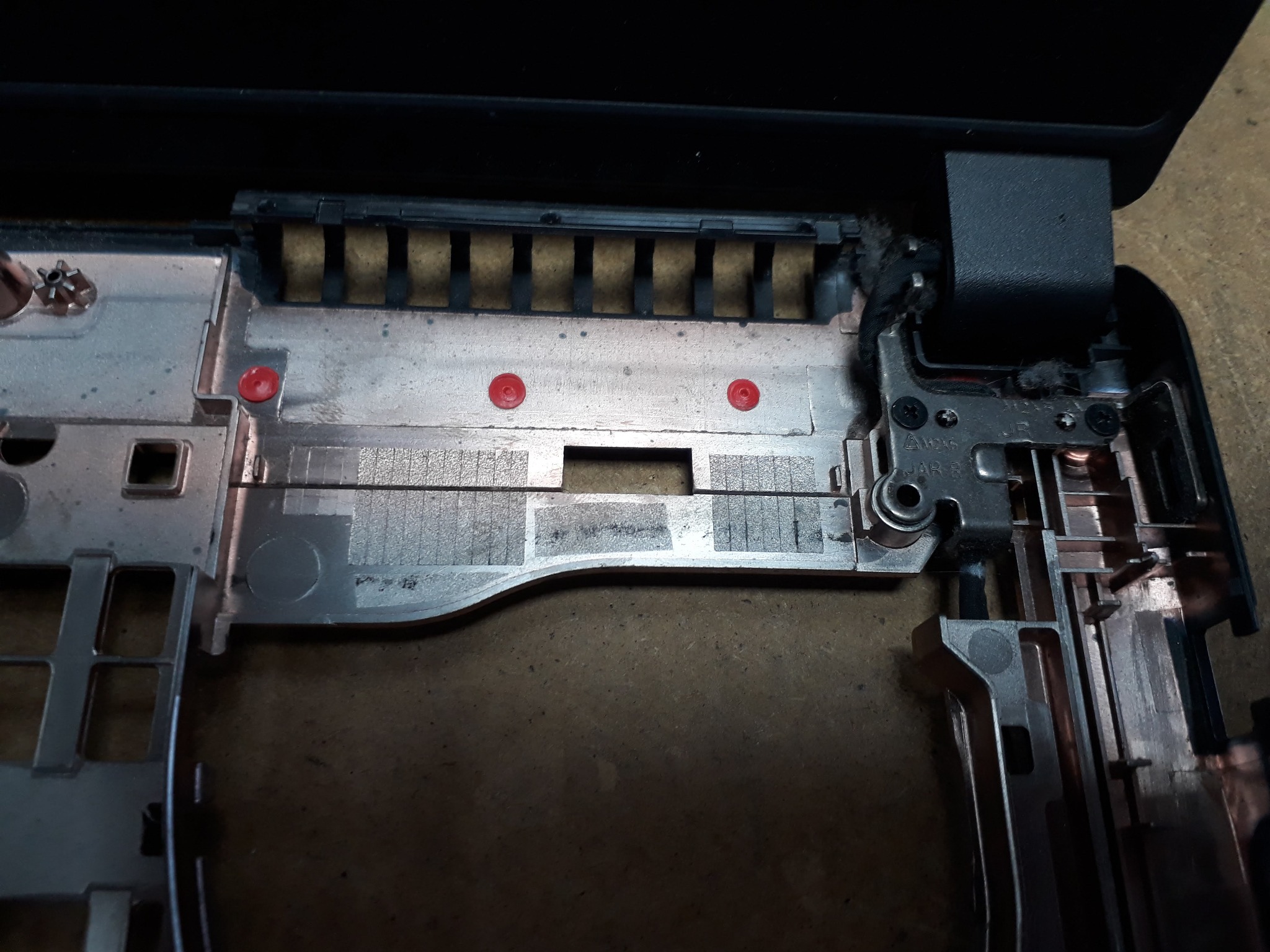 Wnętrze otwartego laptopa z widocznymi elementami mocującymi ekran, zawiasami i metalową konstrukcją wewnętrzną, z trzema czerwonymi elementami mocującymi.