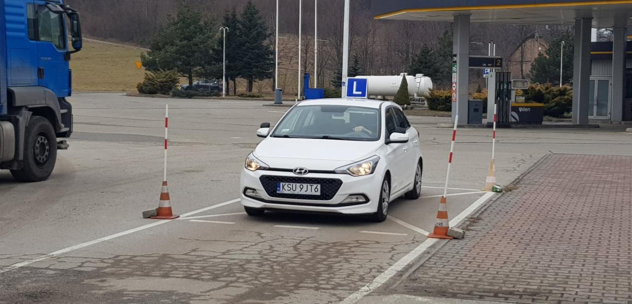 Biały samochód z literą L na dachu, zaparkowany na placu manewrowym z pachołkami, widoczny fragment niebieskiej ciężarówki i stacja benzynowa w tle.