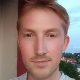 Portret młodego mężczyzny o rudych włosach i brodzie, z delikatnym uśmiechem na tle wieczornego nieba i zabudowy miejskiej.