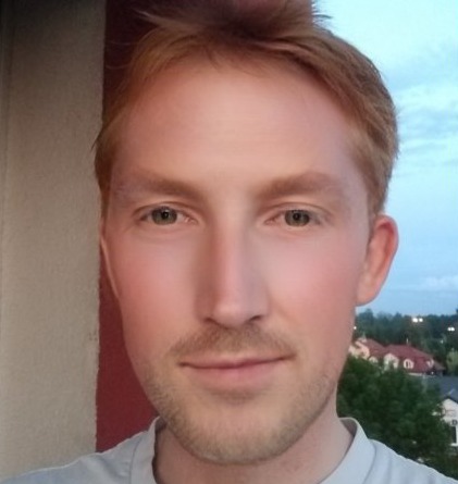 Portret młodego mężczyzny o rudych włosach i brodzie, z delikatnym uśmiechem na tle wieczornego nieba i zabudowy miejskiej.