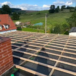 Więźba dachowa z drewnianych łat na czarnej papie, widok z góry na tle zielonych wzgórz i błękitnego nieba z chmurami, obok komina z butelką płynu.