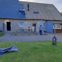 Montaż niebieskiej dachówki na połowie dachu domu jednorodzinnego, widoczna więźba dachowa na niezakończonej części, pracownicy na dachu i mężczyzna z płytą elewacyjną na ziemi.