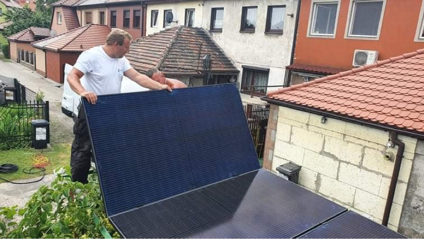 Dwóch mężczyzn instaluje panel słoneczny na dachu domu w zabudowie szeregowej; widoczne okablowanie i fragment dachu sąsiedniego budynku.