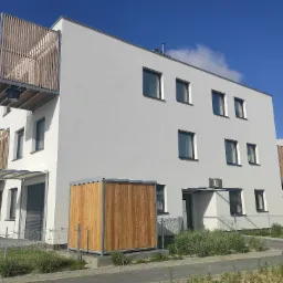 Nowoczesny budynek mieszkalny z białymi ścianami, drewnianymi balkonami i numerem 82 nad wejściem, otoczony zielenią i ogrodzeniem.