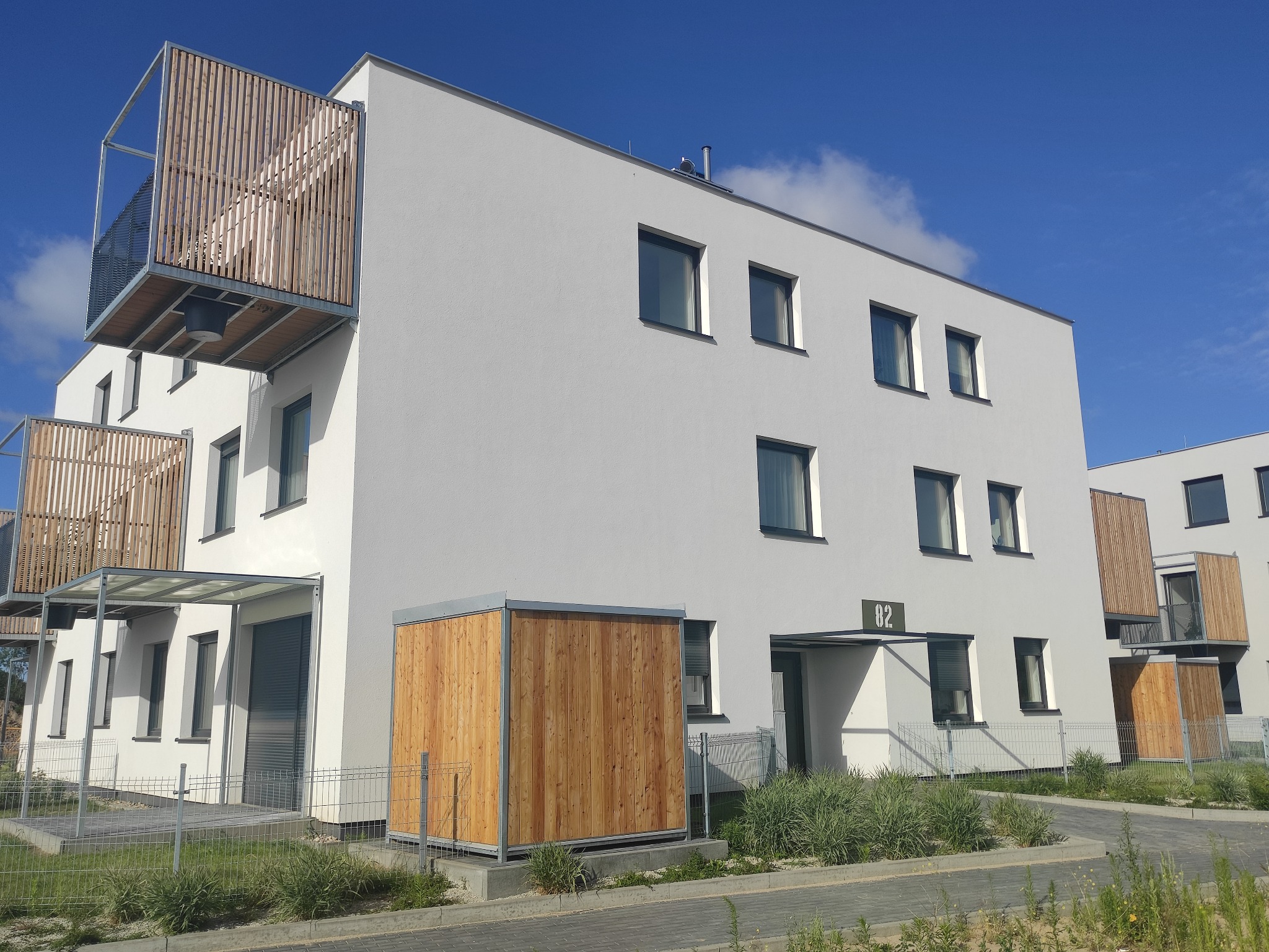 Nowoczesny budynek mieszkalny z białymi ścianami, drewnianymi balkonami i numerem 82 nad wejściem, otoczony zielenią i ogrodzeniem.